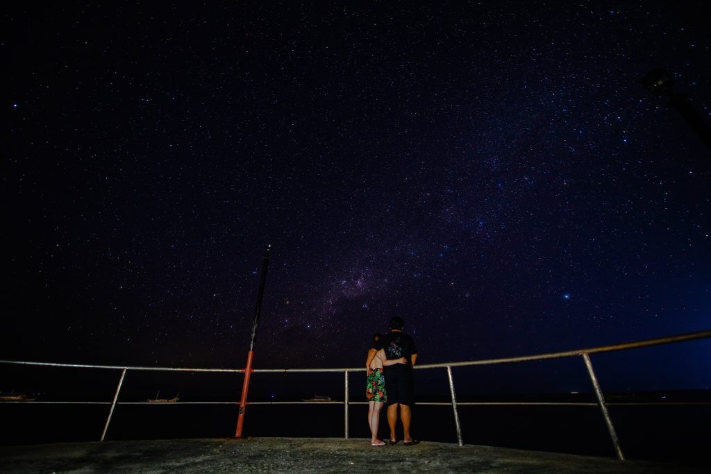 a couple watching the stars in the night sky, standing near a fence